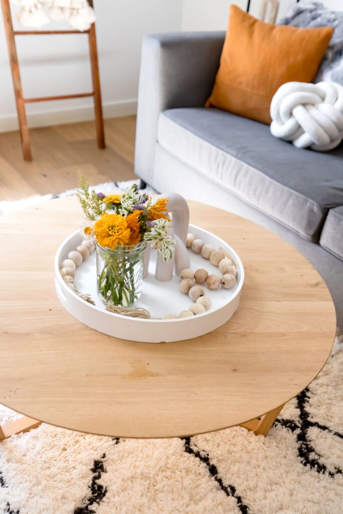 a white tray on a round wooden coffee table. A vase a wood beads are also in the white tray. A blue couch with a fall throw pillow