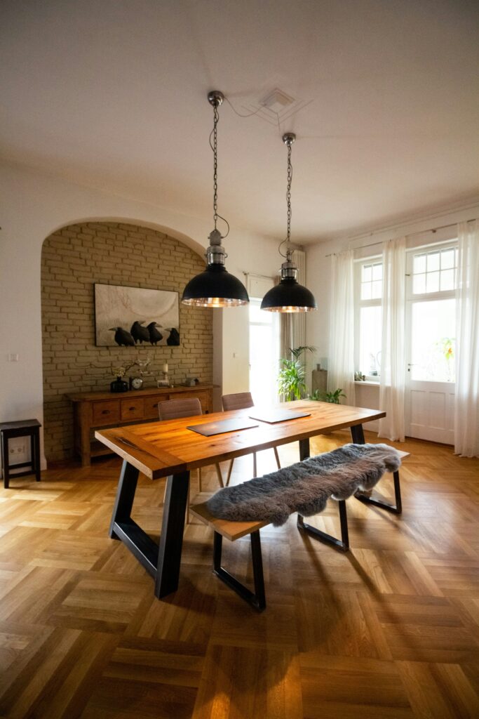 Warm dining room furniture with a large wood dining table, bench seating, black pendant lights, and a wood sideboard against an exposed brick wall.