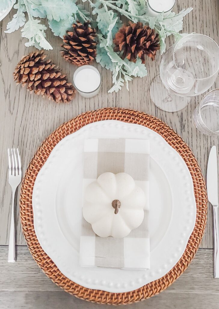White pumpkin on a checked napkin and white plate over a woven charger, with pinecones and pale foliage nearby.