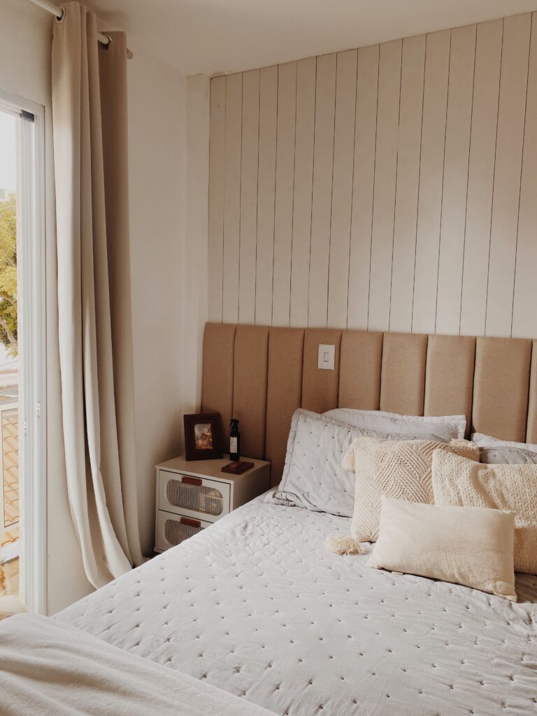 Cozy neutral bedroom with a tall upholstered headboard, layered white bedding, textured pillows, and floor-length curtains.