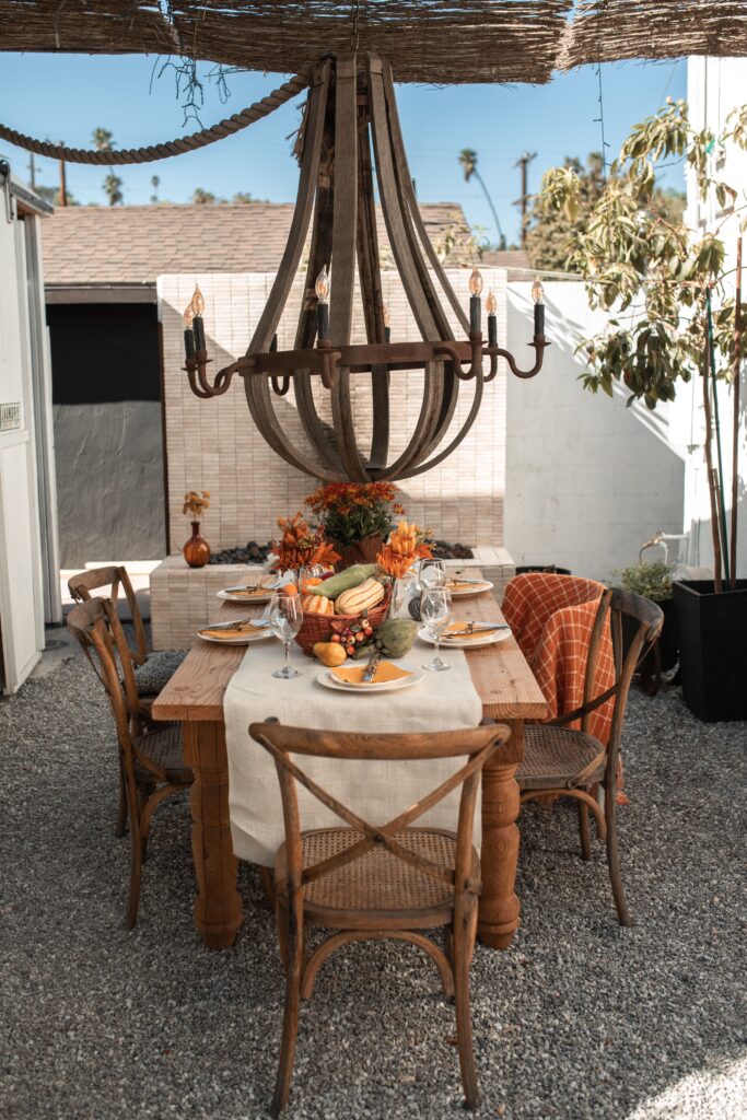 Outdoor wooden dining table with a cream runner, orange flowers, a basket of squash, and an orange checked blanket on a chair.