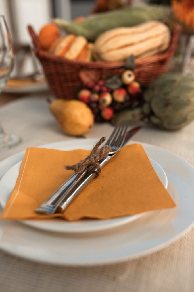 White plates topped with an orange napkin and silverware tied with twine, with a basket of squash behind them.