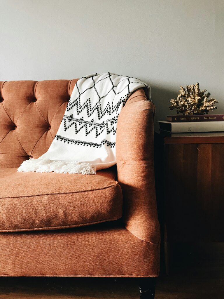 Rust-colored sofa styled with a black and white patterned throw blanket beside a wooden side table with books and decorative coral.